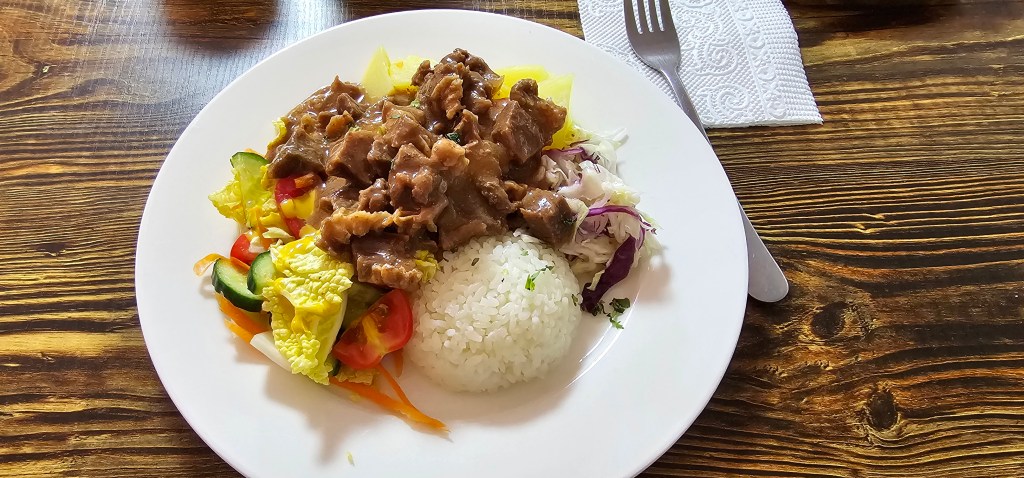 A plate of rice topped with savory beef stew, accompanied by a colorful salad of mixed vegetables, including cucumber, tomato, and cabbage, served on a wooden table.