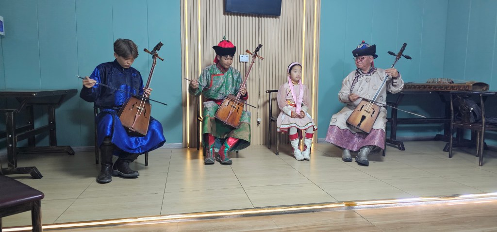 A group of musicians, including an adult male, an adult female, a young girl, and an elder male, are seated indoors playing traditional instruments. The adults are wearing colorful traditional attire, while the girl is dressed in a pink outfit. The setting features a light blue wall and wooden flooring.