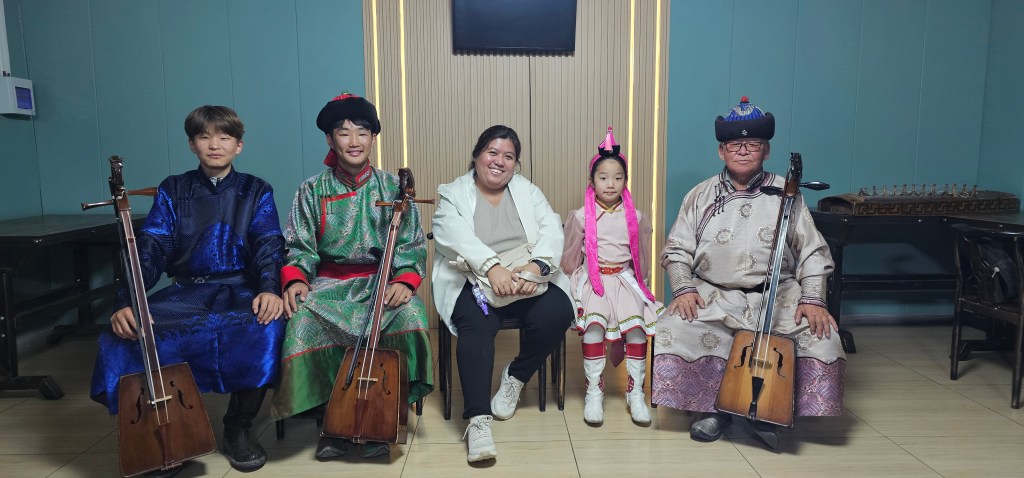 Five musicians posing together, wearing traditional clothing, seated in a room. They hold string instruments, with two children and three adults among them.