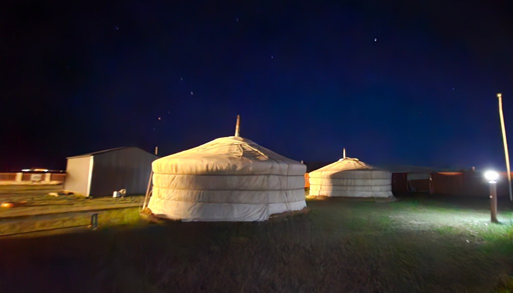 Three traditional yurts illuminated at night, set against a dark sky filled with stars.