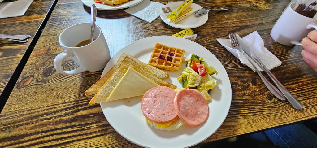 A breakfast plate featuring two slices of toast with cheese, two slices of processed meat, a small salad with vegetables, a waffle topped with jam, and a cup of coffee on a wooden table.