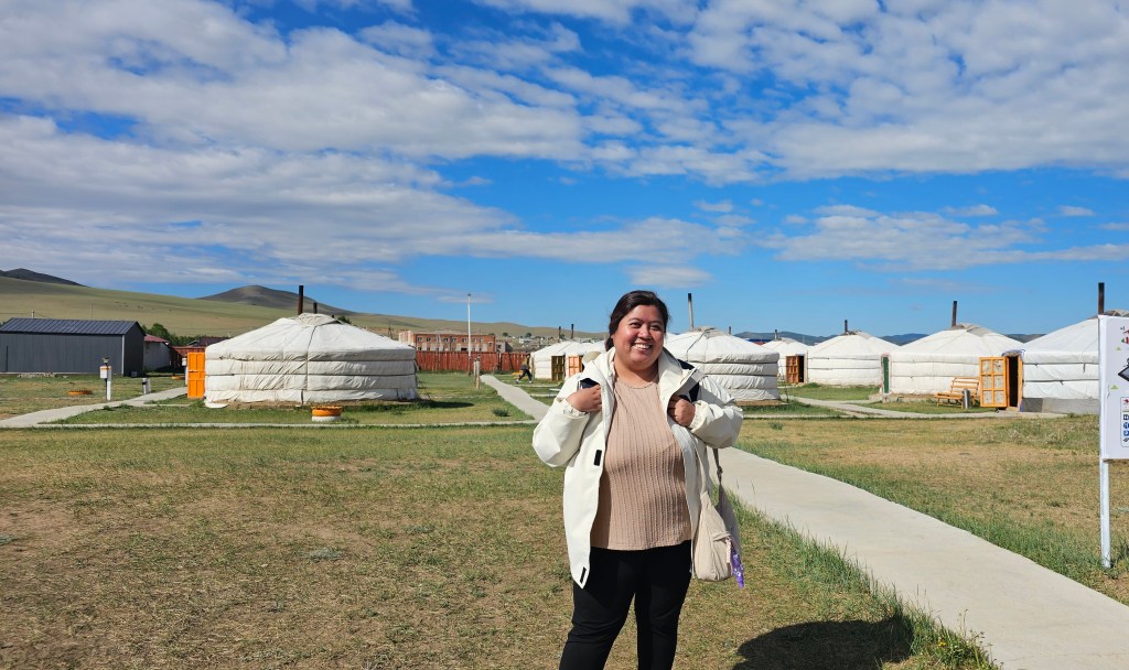 A woman stands smiling in front of traditional yurts in a grassy landscape, under a blue sky with clouds.