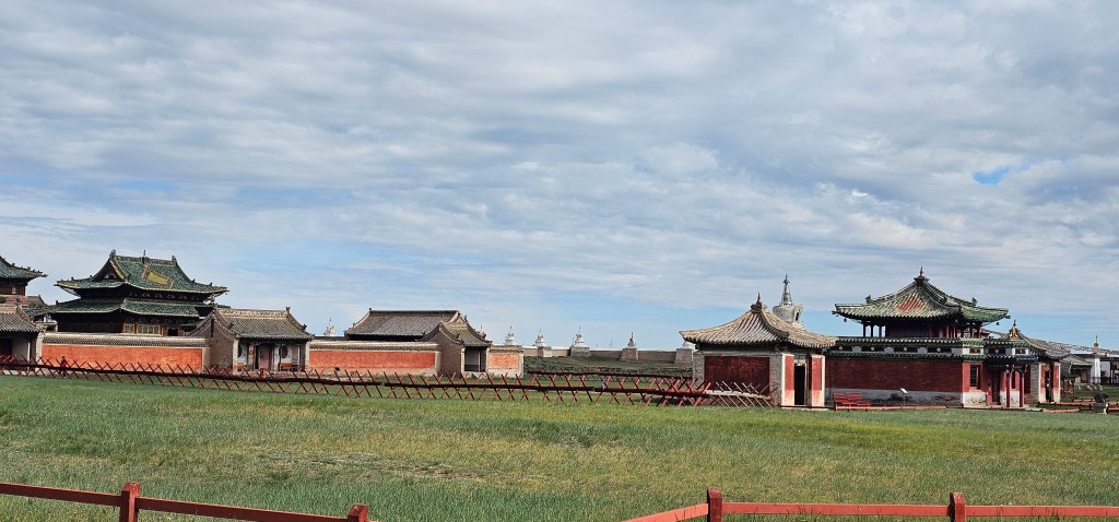 A landscape view of traditional Asian architecture with ornate rooftops and structures, surrounded by grassy fields under a cloudy sky.
