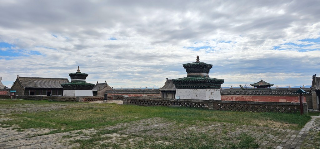 A landscape view of traditional Asian architecture featuring multiple pagoda-like structures surrounded by a grassy area under a cloudy sky.