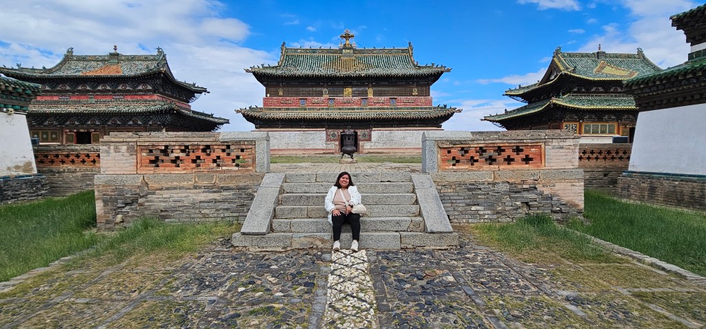 A woman sitting on stone steps in front of traditional Asian architecture with vibrant rooftops, surrounded by green grass and blue skies.