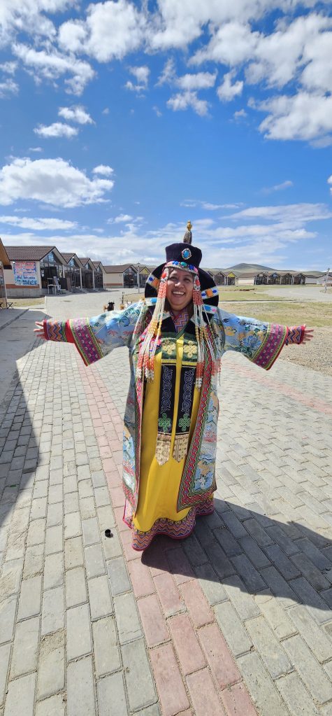 A smiling woman in traditional colorful attire with intricate designs stands outdoors, arms open wide, against a backdrop of blue skies and scattered clouds.