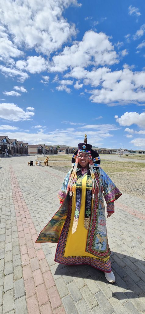 A person wearing traditional colorful attire with intricate patterns, standing outdoors against a clear blue sky with white clouds. In the background, wooden structures and a few dogs can be seen on a paved path.