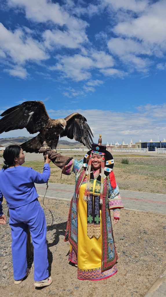 A woman in colorful traditional attire holds a large eagle on her arm while another person in casual clothing assists her, set against a backdrop of blue skies and a grassy landscape.