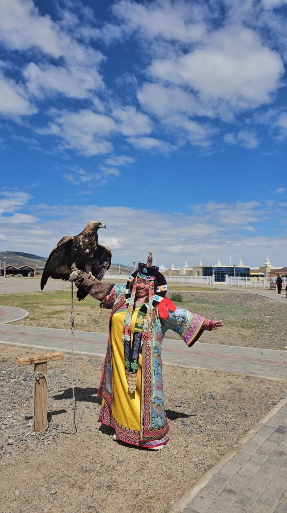A person dressed in colorful traditional clothing poses with a golden eagle perched on their arm, under a blue sky with scattered clouds.