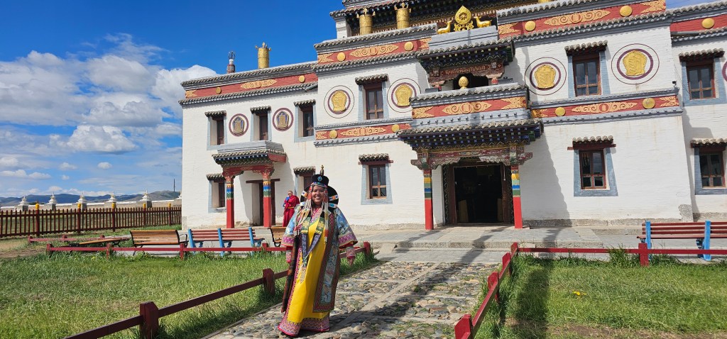 A woman dressed in traditional attire stands in front of a colorful Tibetan-style building with intricate decorations, set against a backdrop of blue skies and fluffy clouds.