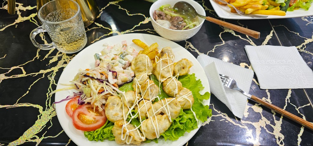 A plated meal featuring fried dumplings drizzled with mayonnaise, served with a side of coleslaw, tomatoes, and lettuce. A glass of beverage and a bowl of soup are also visible in the background.