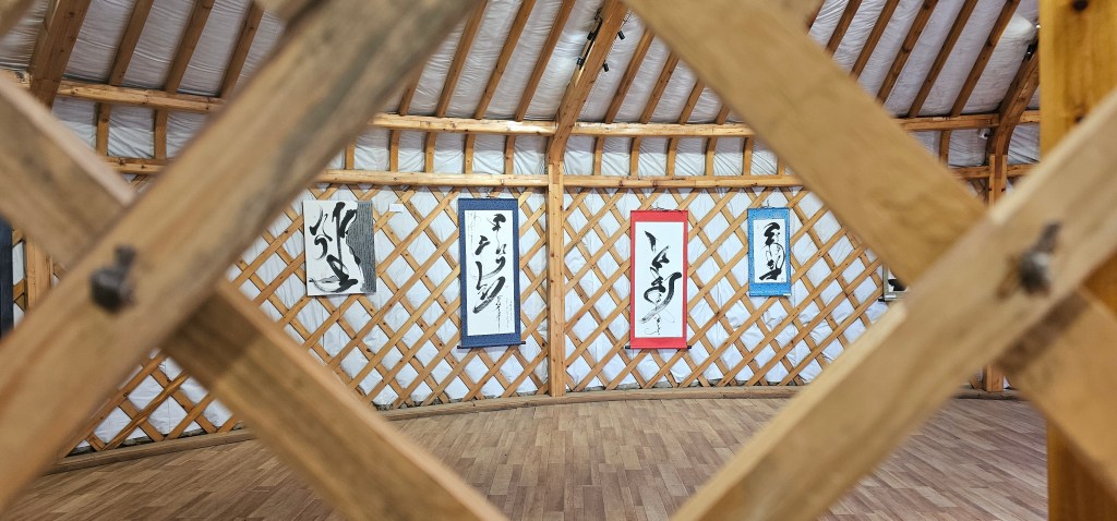 Interior of a wooden yurt with decorative calligraphy scrolls hanging on the walls, viewed through a lattice framework.