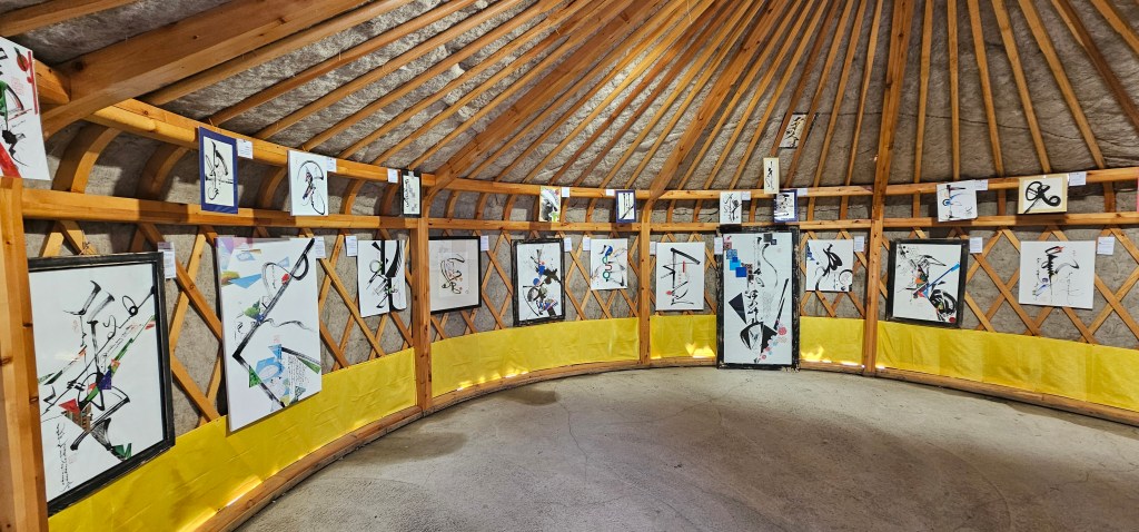 Interior view of a circular room with wooden beams, showcasing various abstract paintings displayed on the walls, with a yellow lower border.