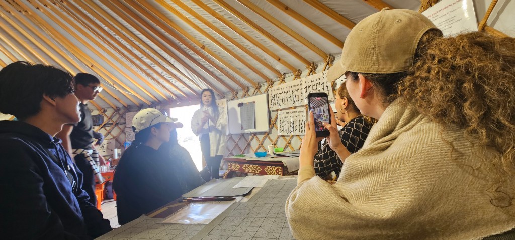 A group of people seated inside a traditional yurt, listening to a presenter. One individual is taking a photo, while others are focused on the speaker and a display on the wall.