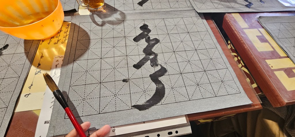 A hand holding a brush above a sheet of gray paper with black ink strokes, surrounded by trays and a drink. The paper has a printed grid design for calligraphy practice.
