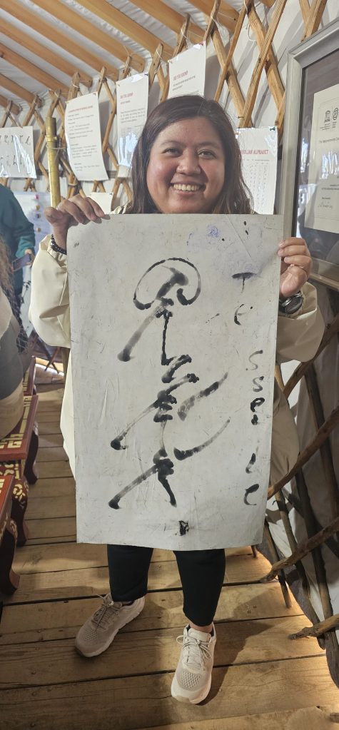 A smiling woman holds a large piece of paper with black calligraphy in a cozy, decorated indoor space.