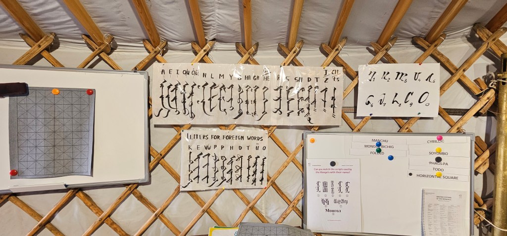 Interior of a yurt displaying educational materials on a wall, including letters for foreign words and various scripts, with a diagram on a whiteboard.