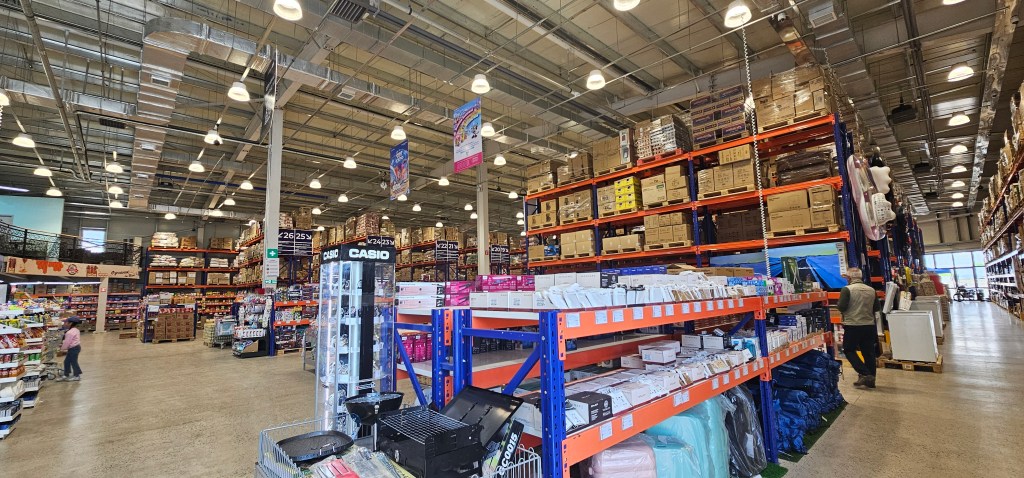 A wide interior view of a retail warehouse showcasing tall shelves stacked with various products, including boxes and household items. The space is brightly lit with ceiling lights, and shoppers can be seen browsing the aisles.