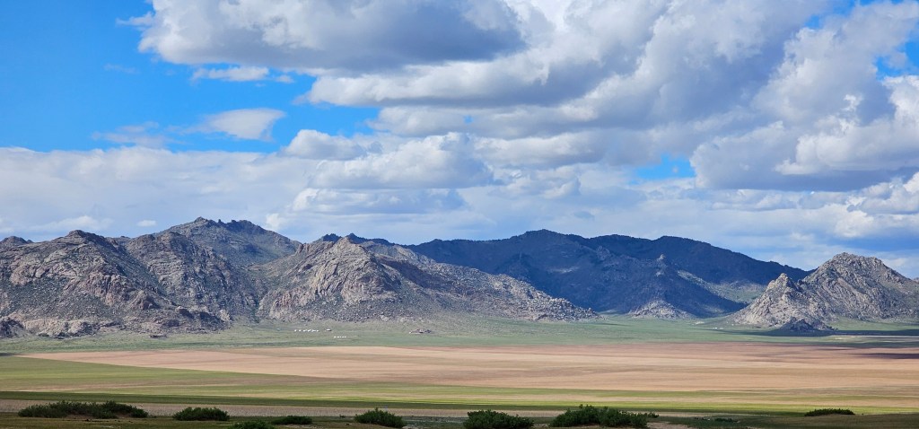 A panoramic view of rugged mountains under a partly cloudy sky, with expansive plains featuring varying shades of green and brown in the foreground.