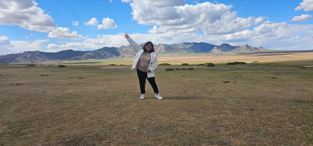 A person standing on a grassy landscape with mountains in the background, smiling and waving. The sky is partly cloudy with blue patches.