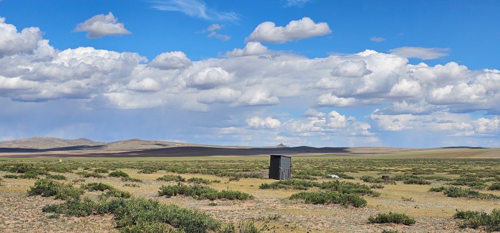 A vast landscape featuring rolling hills, sparse vegetation, and a solitary wooden structure, under a blue sky filled with fluffy white clouds.