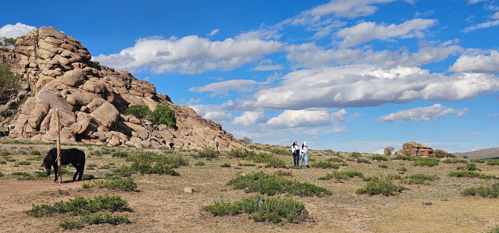 A scenic landscape featuring a black horse grazing on grass, large rocky formations in the background, and two people walking together under a blue sky with scattered clouds.