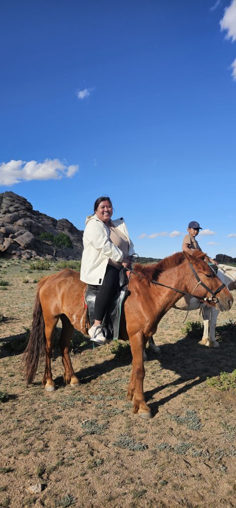 A person sitting on a brown horse with a smiling expression, wearing a white jacket, against a backdrop of blue sky and rocky terrain. Another person is visible on a separate horse in the background.