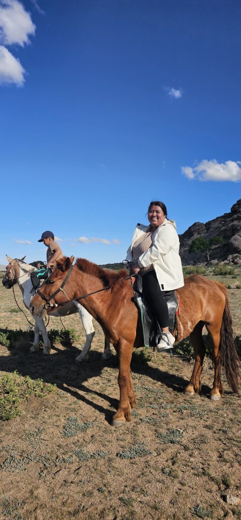 A woman riding a brown horse in a scenic outdoor setting with clear blue skies. A person on another horse is visible in the background.