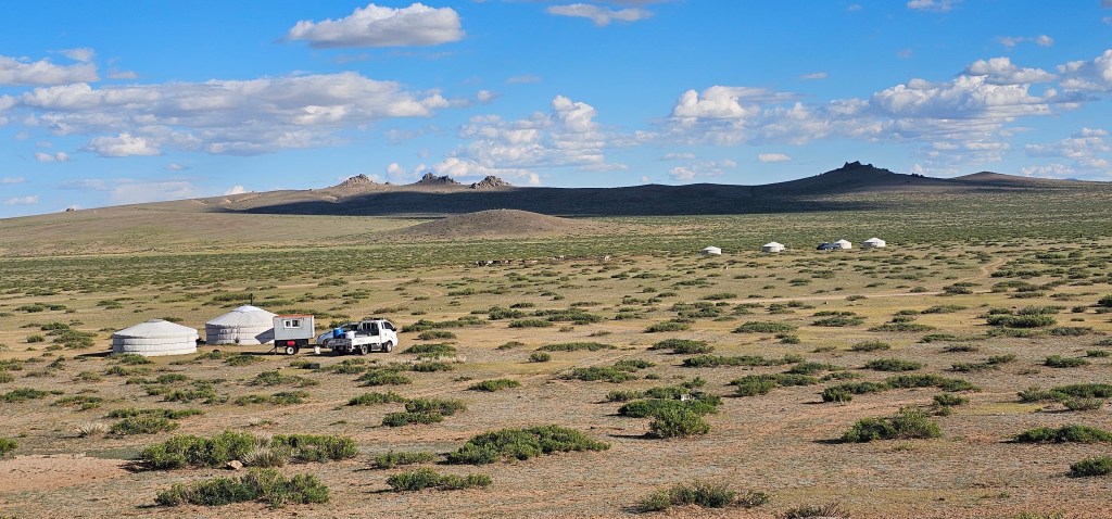 A wide landscape featuring several white yurts and a parked vehicle in a grassy area with distant hills and a blue sky dotted with clouds.