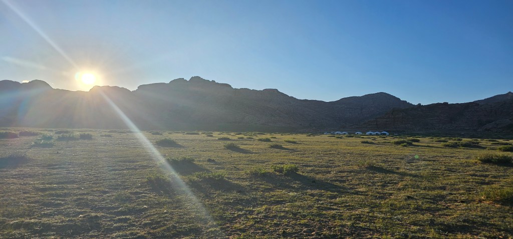 A sunrise over a rocky landscape with grassy fields and distant vehicles visible.