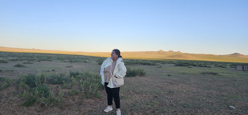 A person standing in a vast open landscape with green vegetation and gentle hills under a clear blue sky.