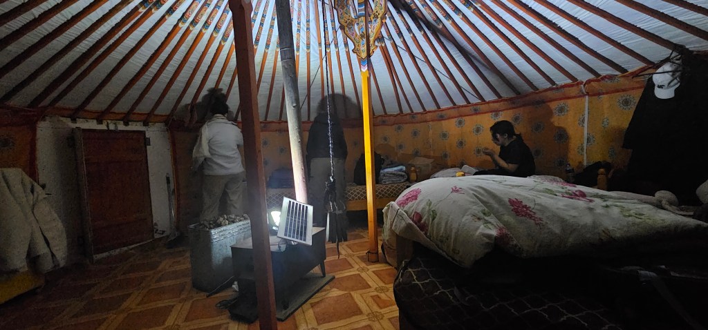 Interior of a cozy yurt with a wooden ceiling, showing two people; one is sitting on a bed with a blanket, while the other is standing near a door.