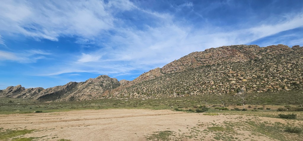 A panoramic view of rugged mountains under a blue sky with wispy clouds, surrounded by green vegetation and an open area in the foreground.