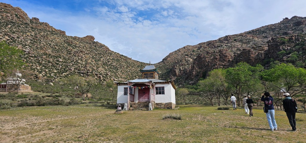 A scenic view of a traditional white building surrounded by lush greenery and rocky mountains, with several people walking towards it.