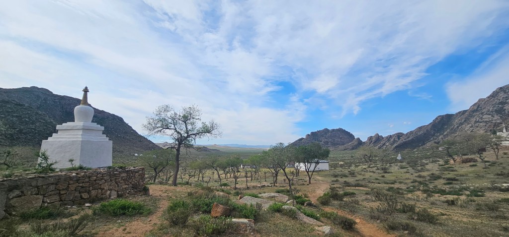 Scenic landscape featuring a white stupa with a golden spire, surrounded by rocky hills and sparse vegetation under a partly cloudy sky.