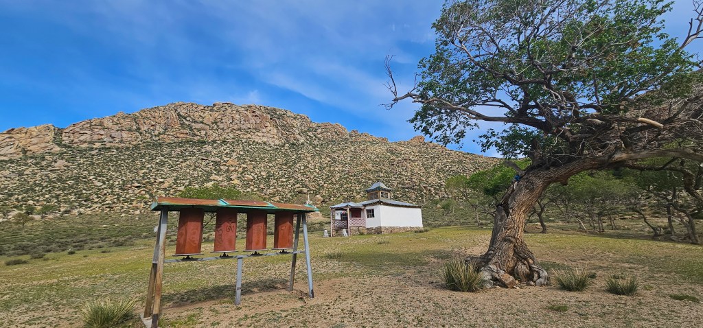 A scenic landscape featuring a hillside of rocky terrain, a small white building with a blue roof, and a wooden structure resembling a beehive in the foreground, along with a large tree to the right.