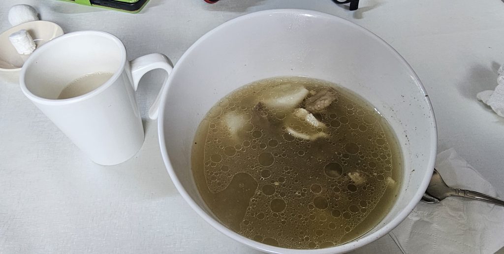 A large bowl of broth with pieces of meat and floating dumplings, accompanied by a white mug of beverage on a table.