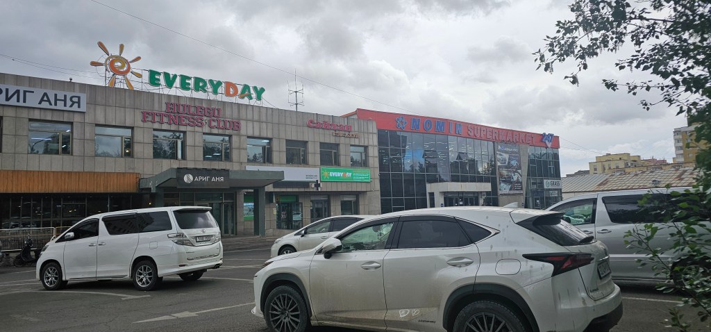 Exterior view of a shopping area with the 'NOMIN SUPERMARKET' and 'EVERY DAY' signage, along with 'HULEGU FITNESS CLUB'. Multiple parked cars are visible in the foreground.