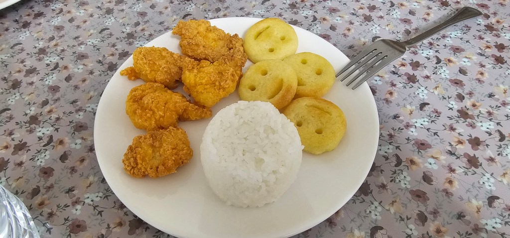 A plate containing fried chicken pieces, a portion of white rice, and golden round-shaped pastries, on a floral patterned tablecloth.