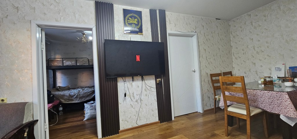 A modern living space featuring a wall-mounted television displaying the Netflix logo, a dining table with chairs, and a door leading to a bedroom with a bunk bed.