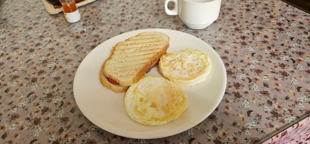 A plate featuring two fried eggs and a slice of toasted bread with a layer of butter or spread, accompanied by a cup of coffee on a floral patterned tablecloth.
