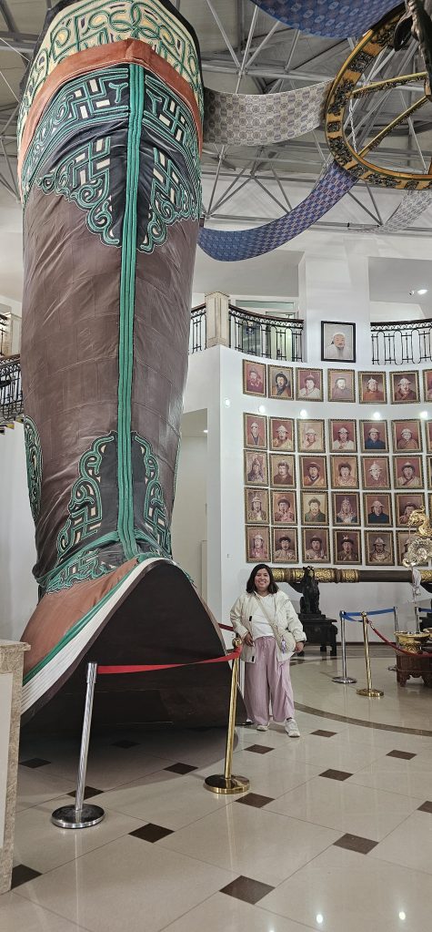 A large decorative boot sculpture displayed indoors, with a person standing beside it for scale. The background features a wall with framed portraits.