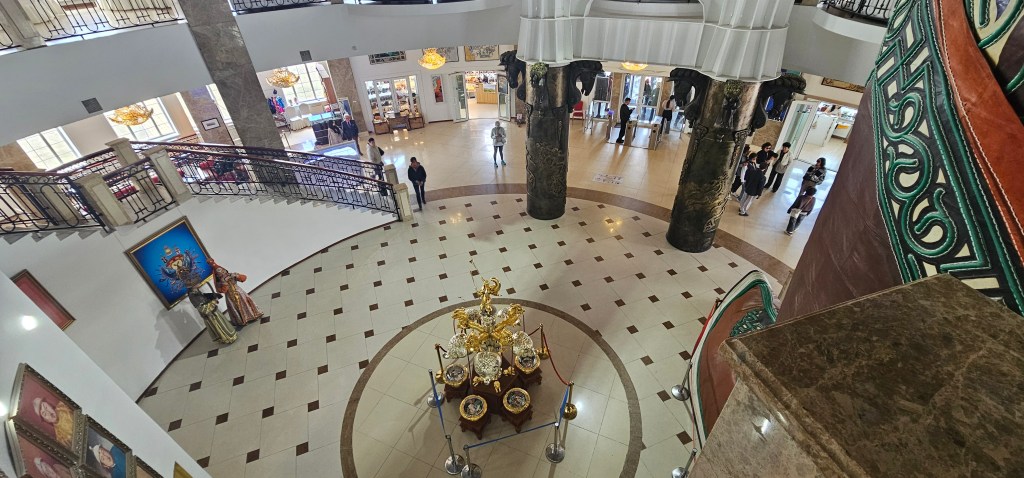 A spacious interior of a building featuring a decorative chandelier, a circular patterned floor, and a central display of ornate golden artifacts surrounded by visitors.