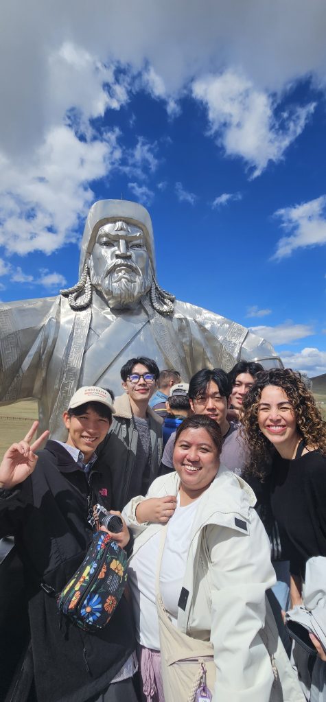 Group of people posing in front of a large silver statue of Genghis Khan under a blue sky with clouds.