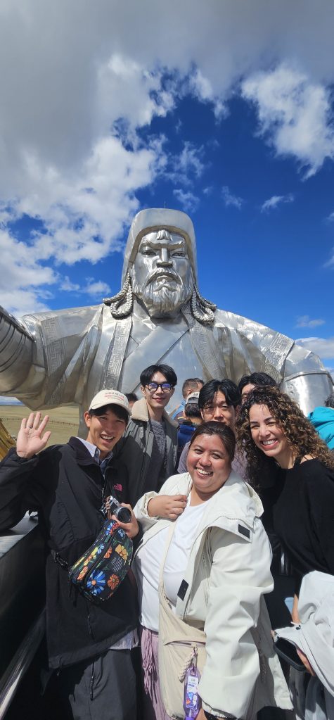 A group of six people smiling and posing in front of a large silver statue of a historical figure, under a partly cloudy blue sky.