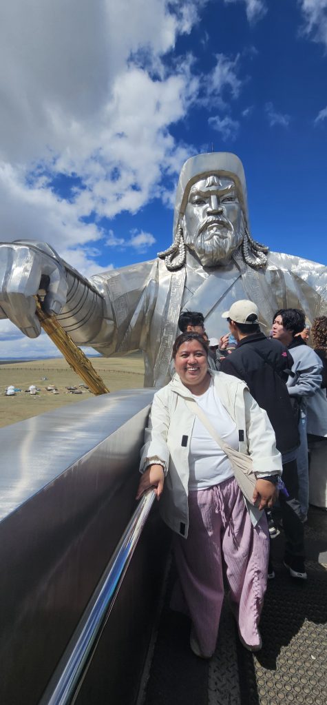 A woman poses in front of a large silver statue of Genghis Khan, with a blue sky and clouds in the background, while other visitors gather nearby.