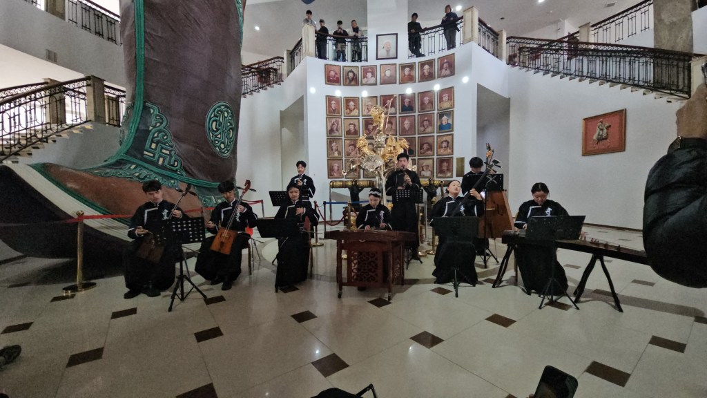 A group of musicians performing in a cultural setting, with a large decorative boot structure in the background and portraits displayed on the walls.