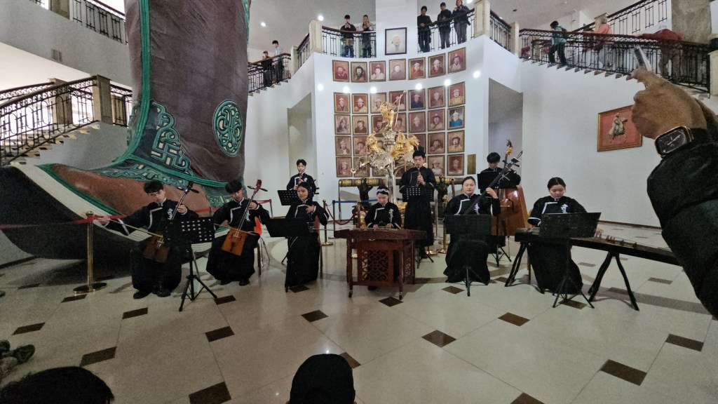 A traditional musical performance in a hall featuring a large decorative boot sculpture, with musicians playing various instruments, including string and percussion, while an audience observes.