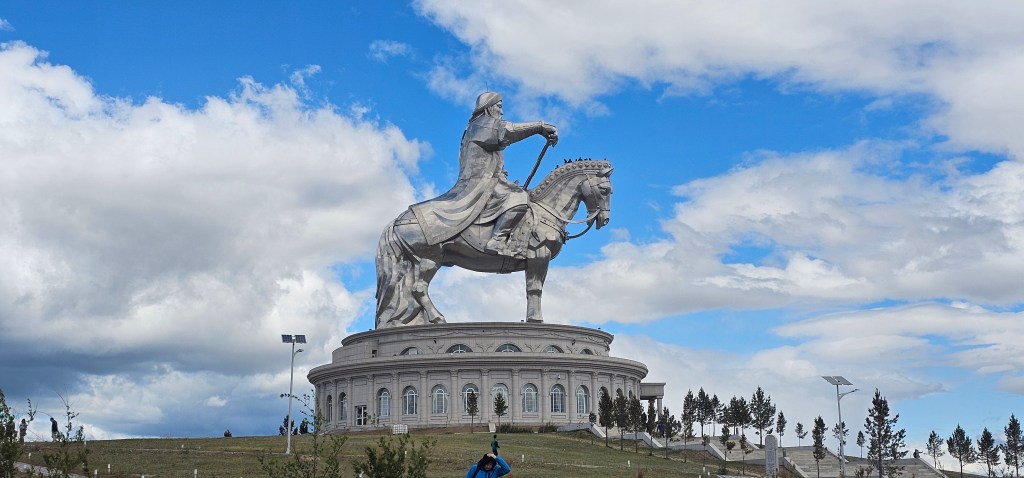 A large statue of Genghis Khan on horseback, situated on a pedestal with a scenic landscape under a blue sky with scattered clouds.