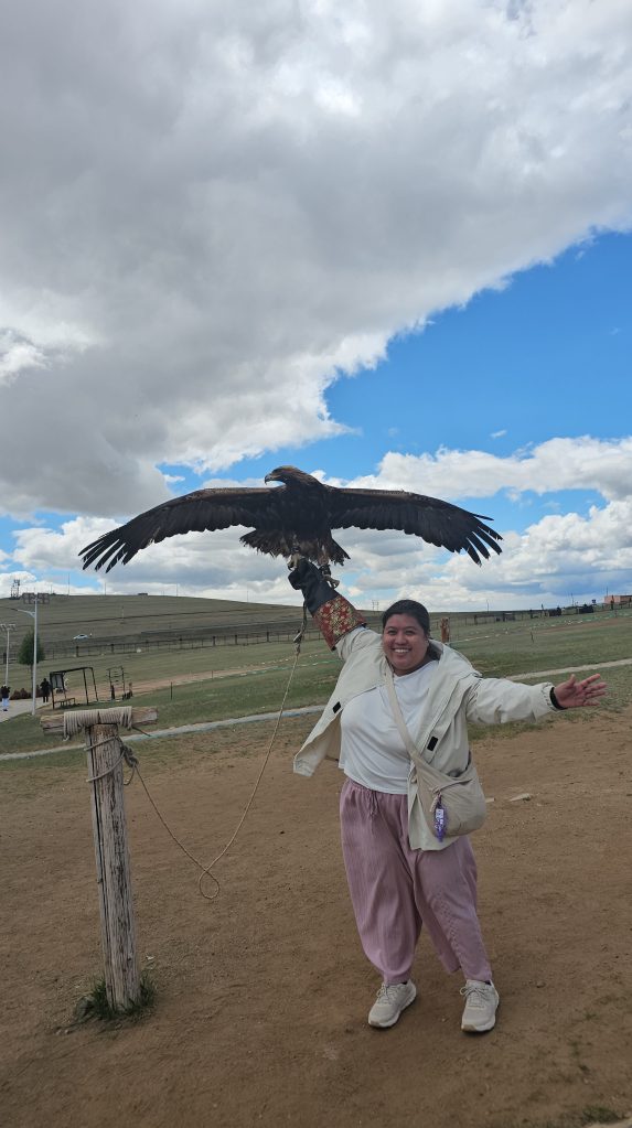 A person stands joyfully with arms outstretched, holding a large eagle on their arm against a backdrop of blue sky and fluffy clouds.